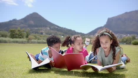 biracial children read books on sunny grass field with mountains