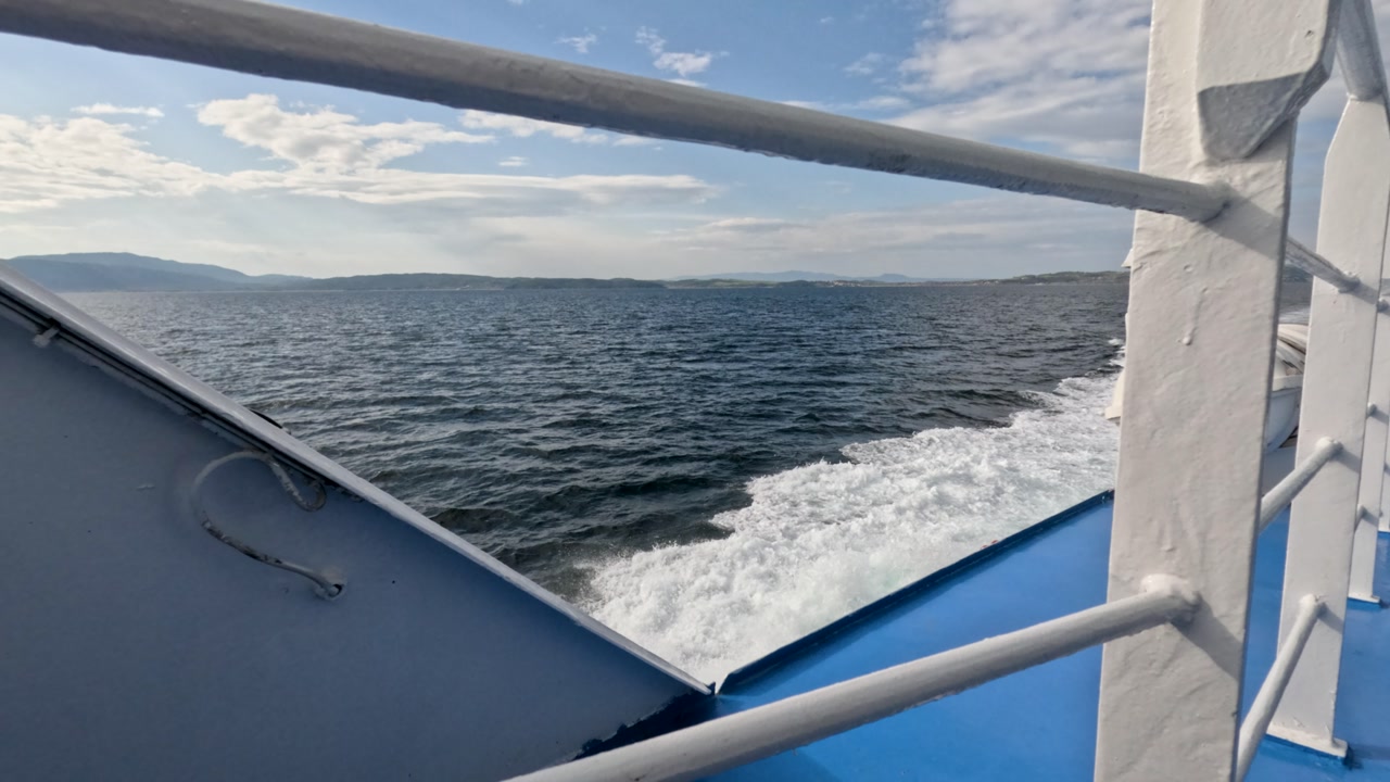 On a ferry trip over the ocean, viewers can see the waves moving and land far away. The sun shines bright and the sky is clear, creating a lovely day for sailing.