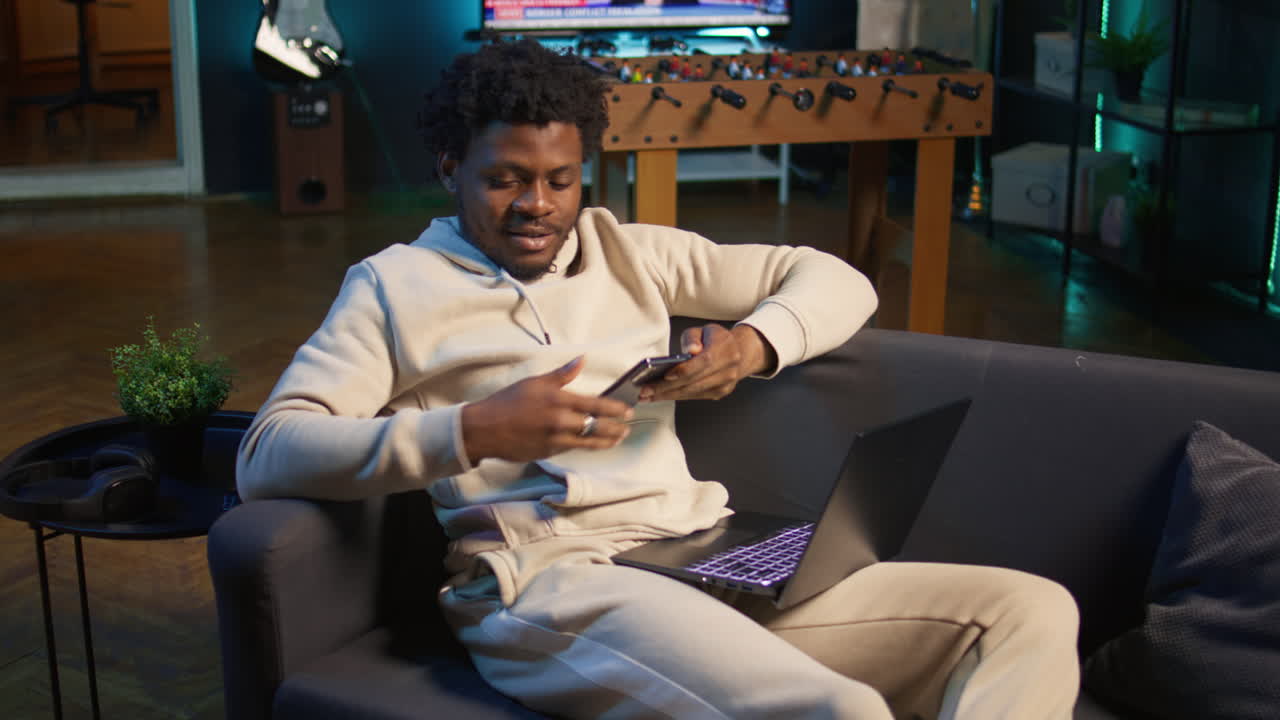 Happy self employed person enjoying a break with his mobile phone, texting friends on his smartphone with a laptop nearby. Cozy living room showcase work and leisure in remote lifestyle. Camera B.
