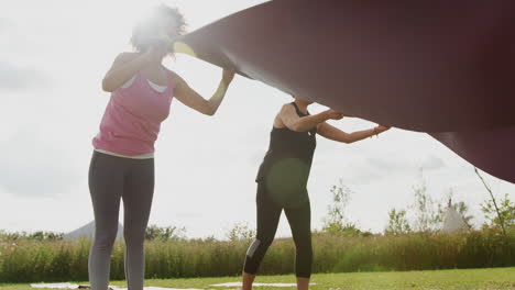 Dos-Amigas-Maduras-Tendiendo-Colchonetas-De-Ejercicio-Sobre-El-Césped-En-Un-Retiro-De-Yoga-Al-Aire-Libre