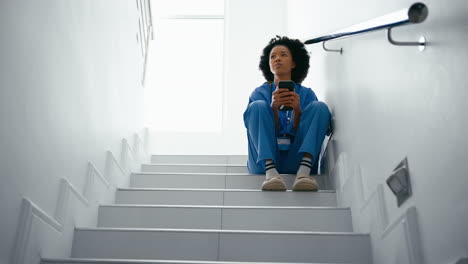 stressed female nurse wearing scrubs browsing on mobile phone sitting on stairs in hospital