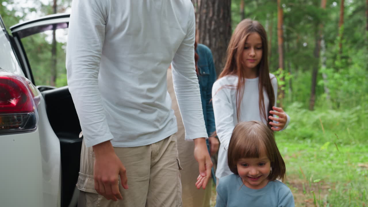 Side view of boy gently lifting his sister down from white car while mother stands nearby smiling warmly in lush forest setting, surrounded by greenery, and peaceful family travel atmosphere