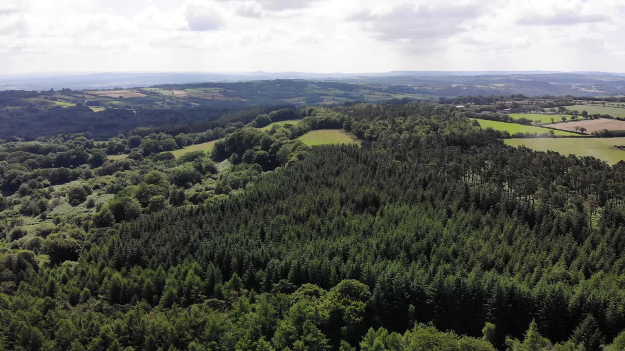 Aerial view of the Blackdown Hills countryside showcasing lush greenery, dense forests, and picturesque open fields under a partly cloudy sky. Pull back Shot