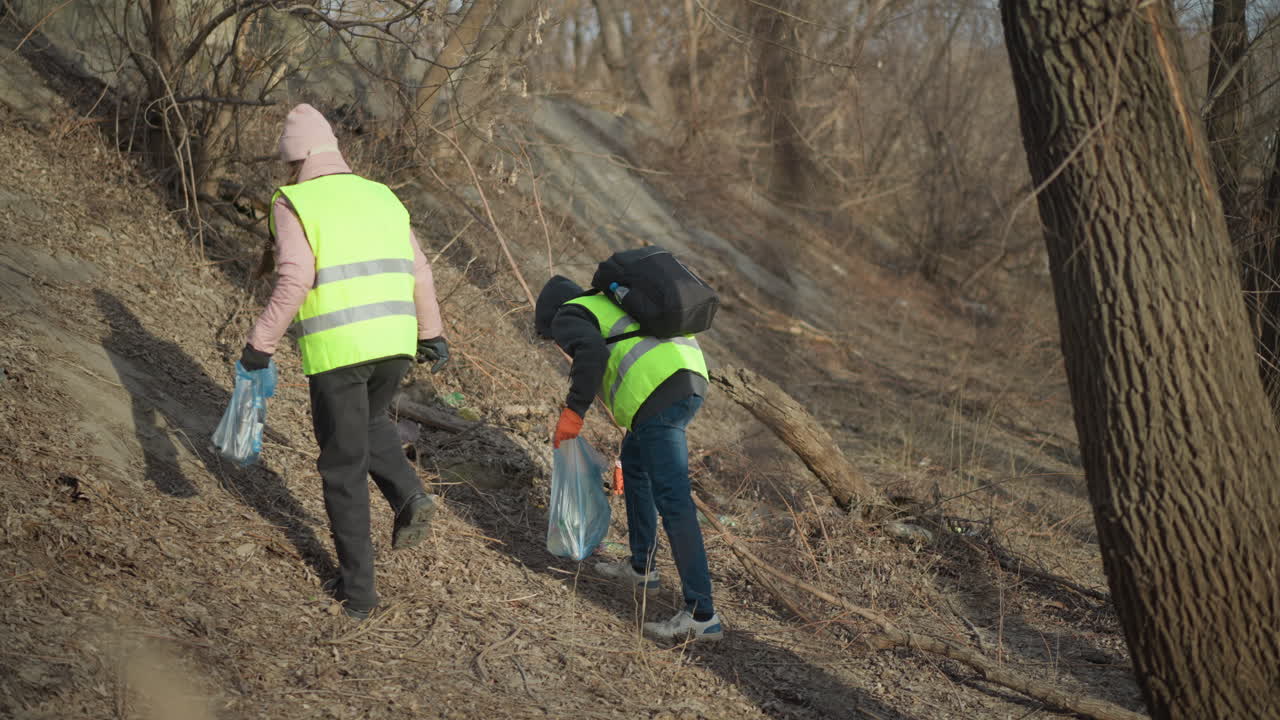 Two volunteers wearing reflective safety vests and gloves carrying bags of collected trash while walking along riverbank during outdoor cleanup, removing litter to protect environment