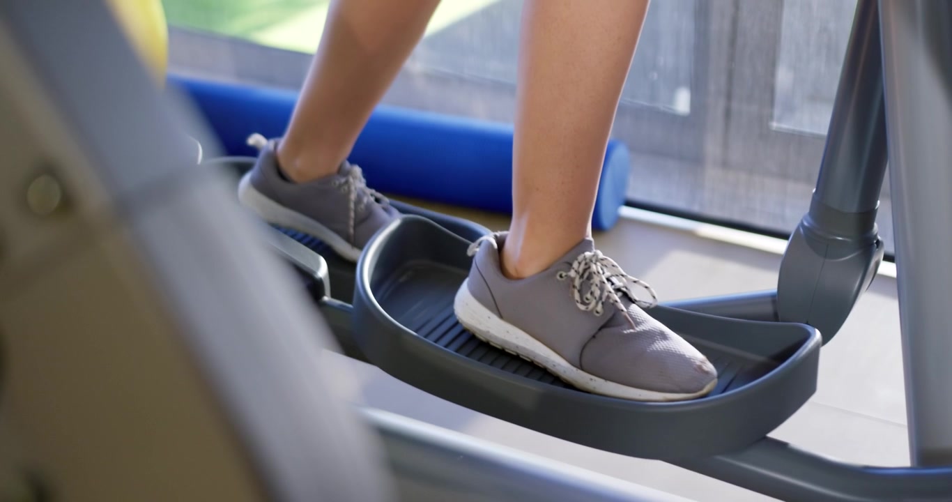 Hombre empujando el pedal derecho, alternando los pies en la elíptica para un entrenamiento cardiovascular junto a la ventana.
