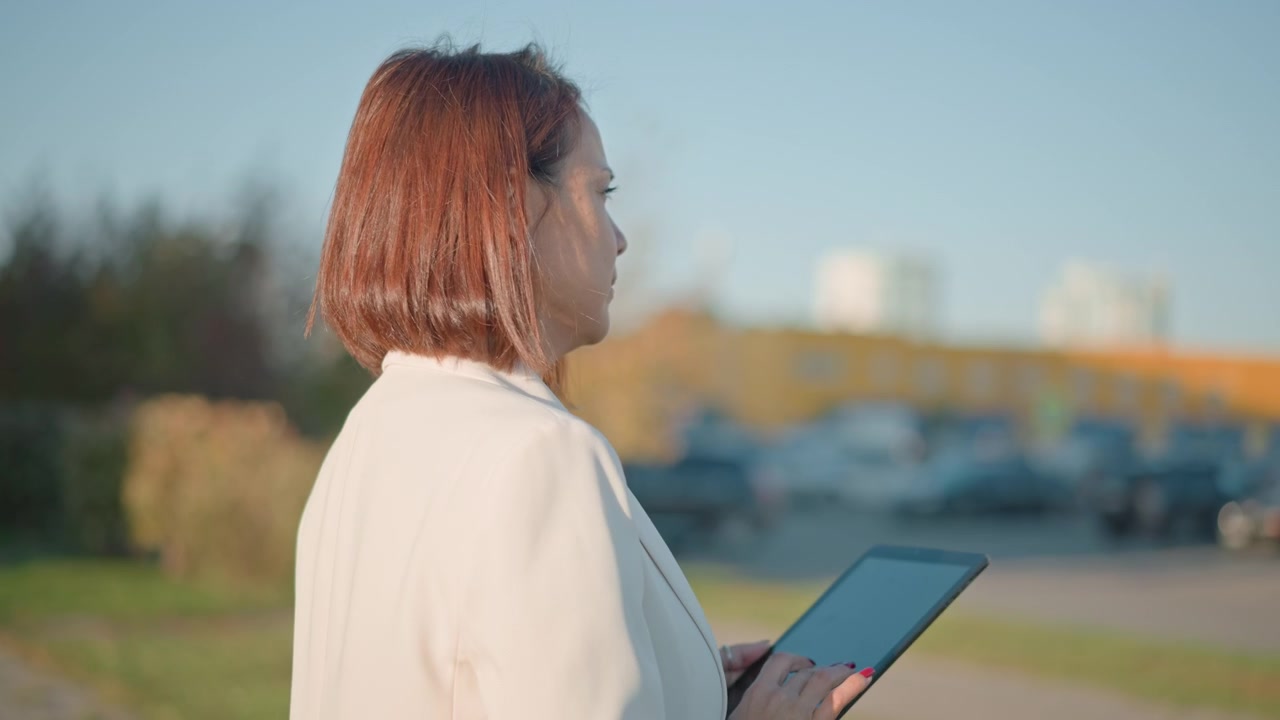 Perfil de mulher de meia-idade em pé no parque, segurando um tablet com as duas mãos enquanto olha para a tela, com uma leve inclinação da cabeça e linguagem corporal calma, demonstrando leitura e interação focadas.