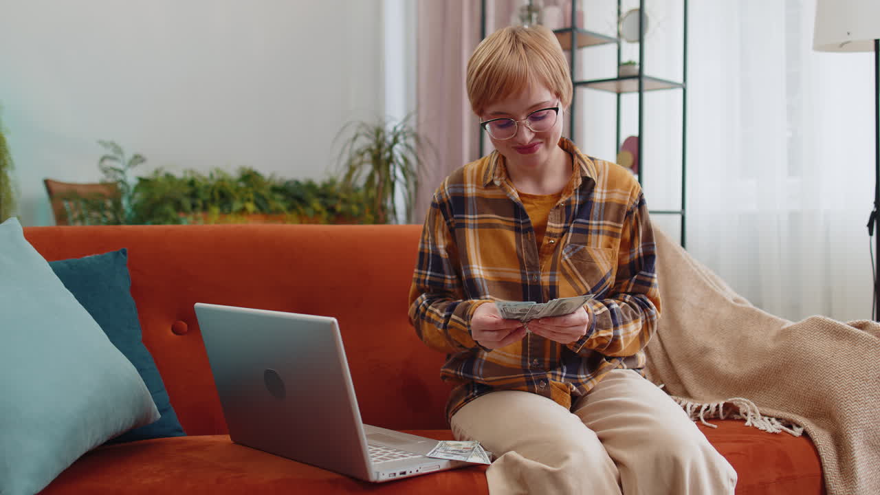 Smiling happy woman counting money cash and use laptop pc calculate domestic income earnings at home