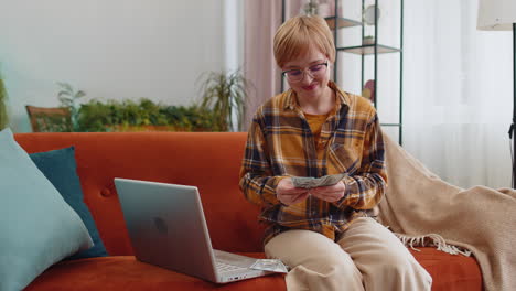 Una-Mujer-Feliz-Y-Sonriente-Contando-Dinero-En-Efectivo-Y-Usando-Una-Computadora-Portátil-Para-Calcular-Los-Ingresos-Domésticos-En-Casa