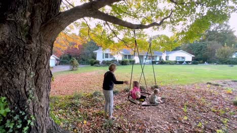 grandmother swings grandkids on a rope swing under a large maple in fall