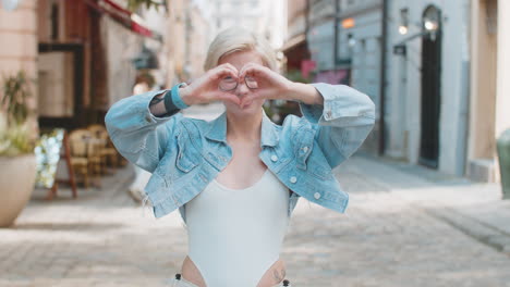 portrait of smiling young woman makes symbol of love showing heart sign to camera on city street