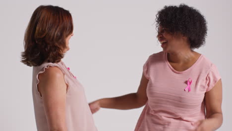 studio portrait of two smiling mature women wearing pink breast cancer awareness ribbons hugging against white background