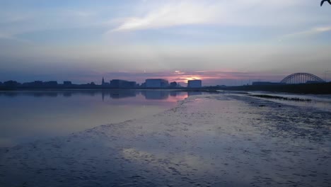 Beeindruckender-Sonnenaufgang-über-Einem-Industriestadtgebiet-Mit-Eiserner-Bogenbrücke,-über-Dem-Flussufer,-Während-Ein-Vogel-Vorbeifliegt,-Zeitlupe---Slomo,-Drohnenantenne