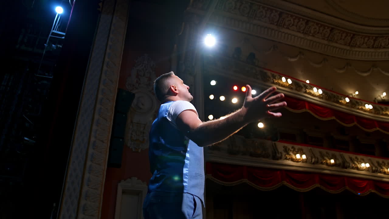 Showman entertains the audience under the light, side view. Big stage. Serious performer speaks from the stage with streched hands.