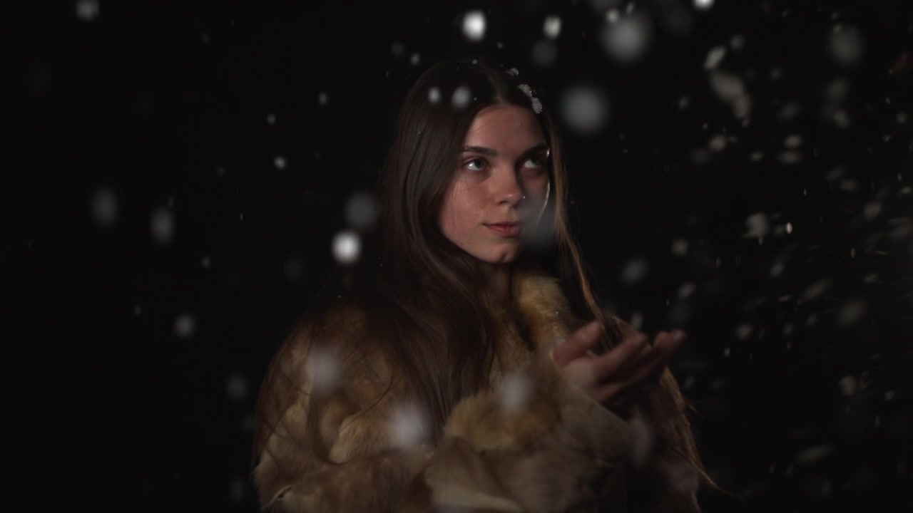 Una donna elegante è mostrata di profilo e parzialmente di schiena sotto un'illuminazione bassa e drammatica, mentre indossa un colletto o un cappotto di pelliccia e particelle leggere fluttuano nell'aria.