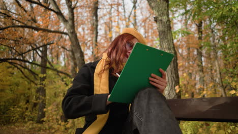 artist thoughtfully sketching on canvas, seated with head tilted to the left on wooden park bench, surrounded by autumn foliage, dressed in stylish yellow beret and scarf