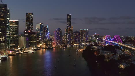 boats in brisbane river at night with beautiful view of illuminated story bridge and skyscrapers in brisbane, queensland - aerial drone, wide shot