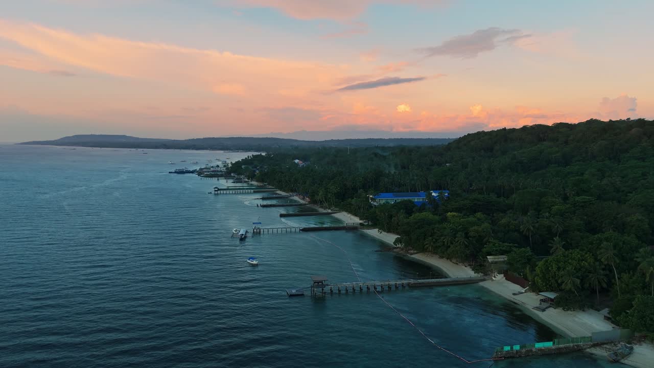 Stunning 4K aerial of a tropical Philippine coastline with sandy beaches, boats, piers, lush forest, and pastel sunset clouds over the horizon