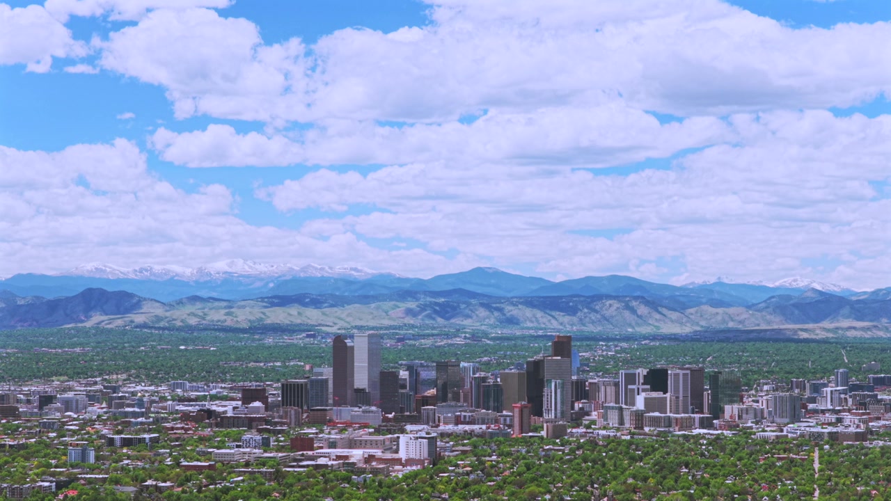 Denver, Colorado: Droneoptagelse af byens skyline, skyskrabere og downtown med de sneklædte Rocky Mountains i Front Range. Forårsmorgen med blå himmel og skyer over byens kvarterer, med en fremadgående panorering opad.