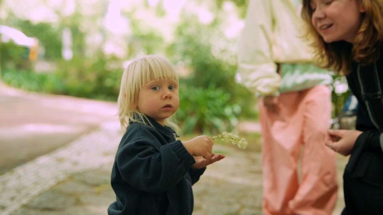 Blonde Toddler Smelling Flowers and Interacting with Adult in Park
