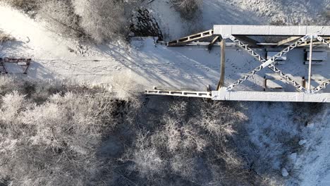 Birds'eye-view-of-an-abandoned-railway-track-on-bridge-over-river-heading-into-forest,-aerial