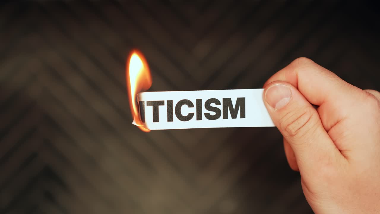 Close-up hand holds a paper strip labeled “CRITICISM” as the edge burns with flame on a dark background. Concept of ignoring negativity, resilience, and mindset change