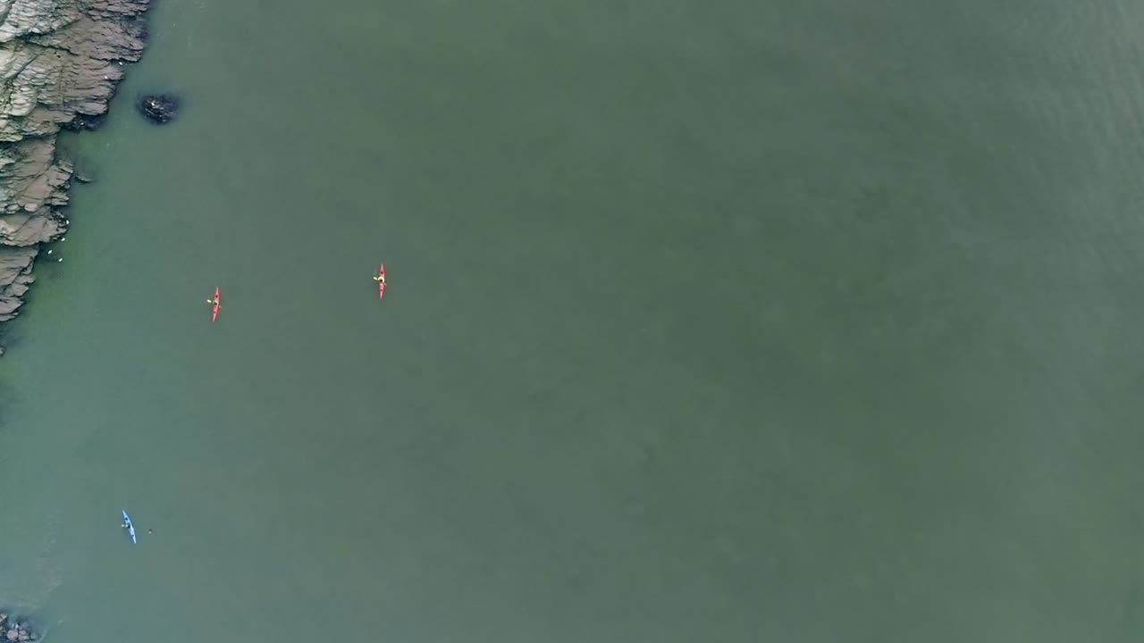 seguimiento aéreo de derecha a izquierda a través de una gran masa de agua turbia con tres kayakistas remando río abajo y una costa rocosa a su lado.