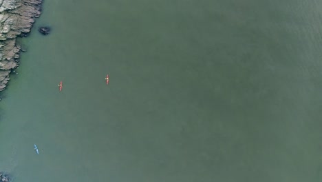 seguimiento aéreo de derecha a izquierda a través de una gran masa de agua turbia con tres kayakistas remando río abajo y una costa rocosa a su lado.