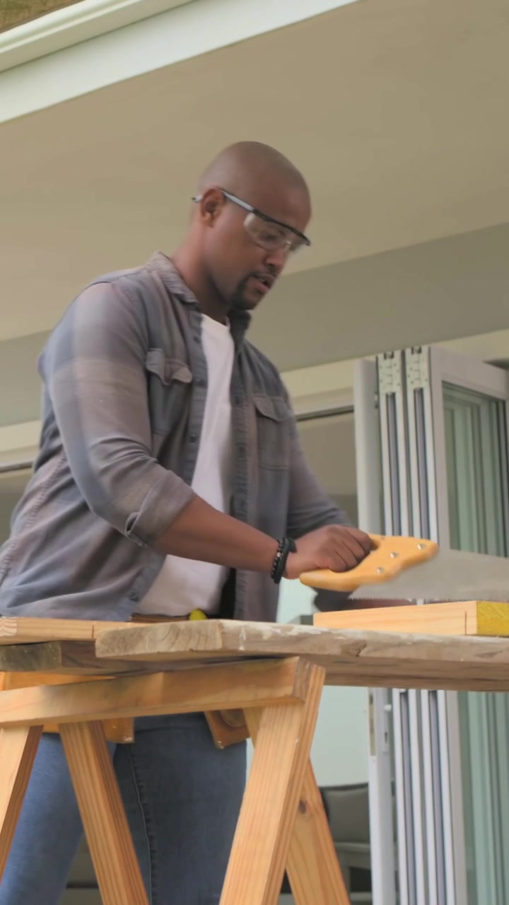 Vertical video: Placing lumber man in grey sawing wood at patio bench prepping boards, copy space
