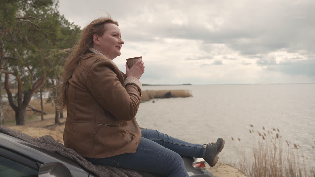 Red Haired Beautiful Woman Sitting On Her Car Next To A Lake, She Drink A Hot Beverage
