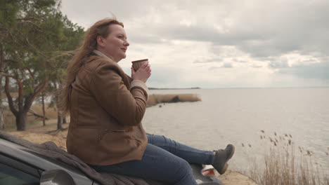 red haired beautiful woman sitting on her car next to a lake, she drink a hot beverage