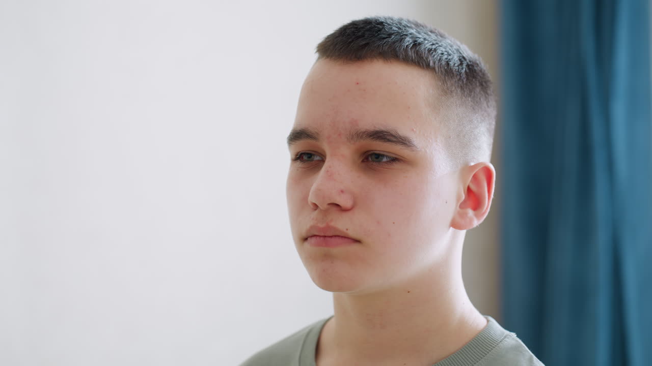 Close up of young boy with calm yet confident expression staring ahead in softly lit room, subtle acne and natural skin texture highlight authentic teenage look in quiet indoor setting