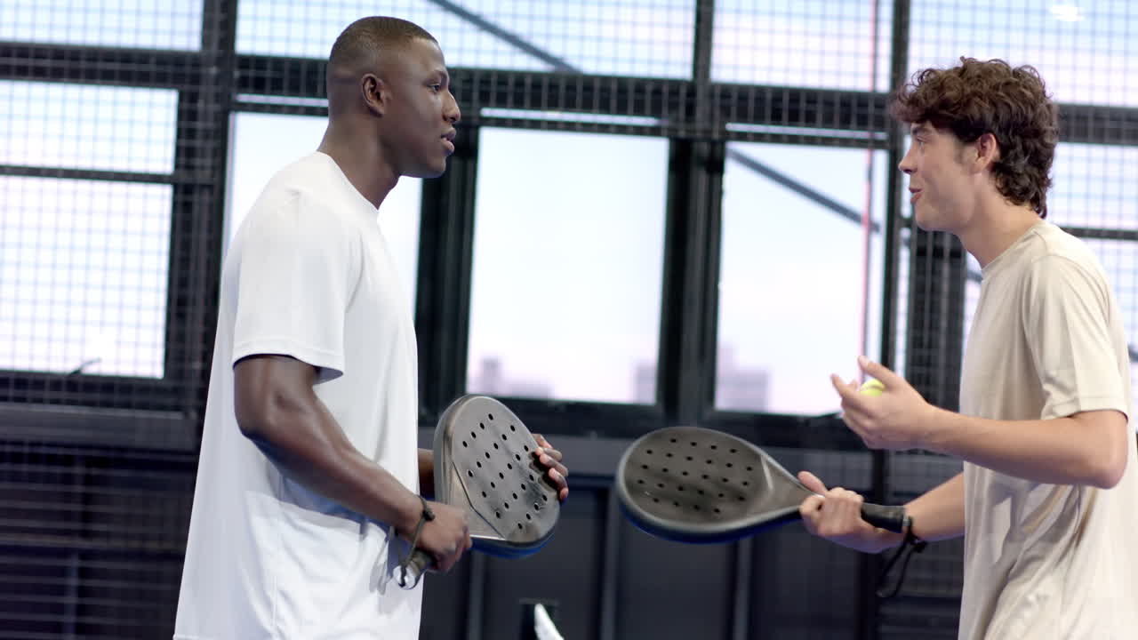 Two diverse men enjoying padel tennis on indoor court, smiling and holding rackets
