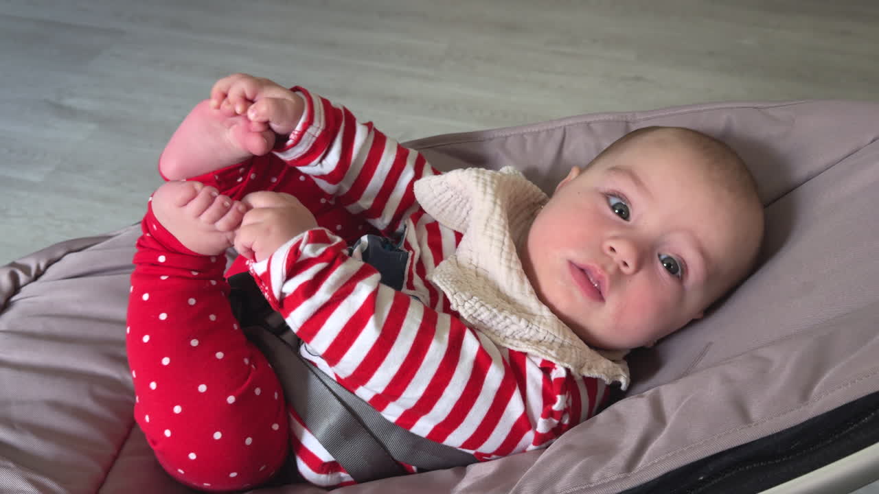 Cute four-month-old baby girl lying on a bouncer, holding her tiny feet playfully. Warm home setting, showcasing early childhood happiness, innocence, and natural playful behavior