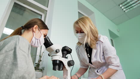 female student in mask looks at microscope attentively. professor helping the student. low angle view.