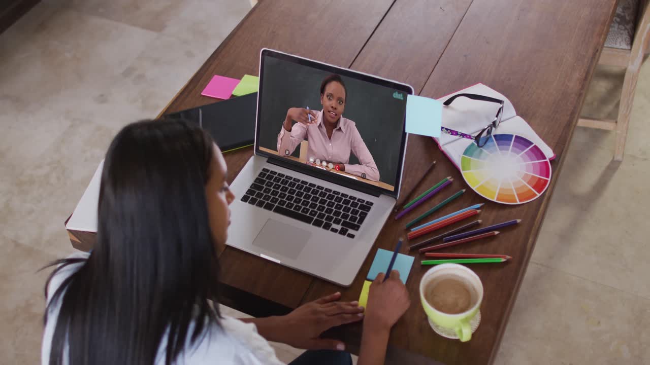 Estudiante caucásica usando una laptop en videollamada con una profesora, tomando apuntes.