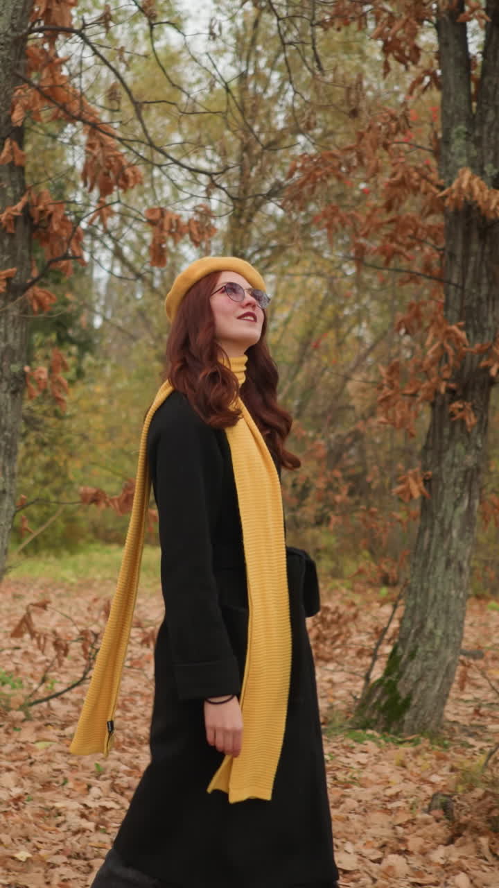 Schöne Dame im Wald spaziert mit einem gelben Barett und passendem Schal, richtet sich mit der linken Hand die Haare zurecht, während sie eine Sonnenbrille trägt