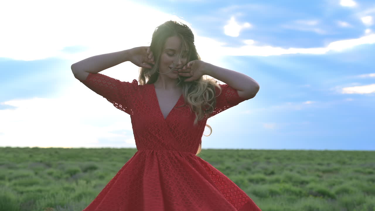 A woman in a red dress walks through a lush green field with the wind in her hair
