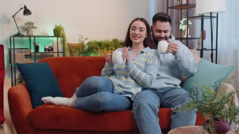 Smiling-family-couple-man-woman-drinking-a-cup-of-coffee-or-herbal-tea-at-home-sofa-in-the-morning