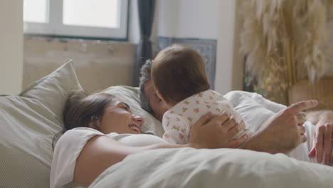 happy couple playing with their cute baby girl and kissing her while lying in bed on sunday morning