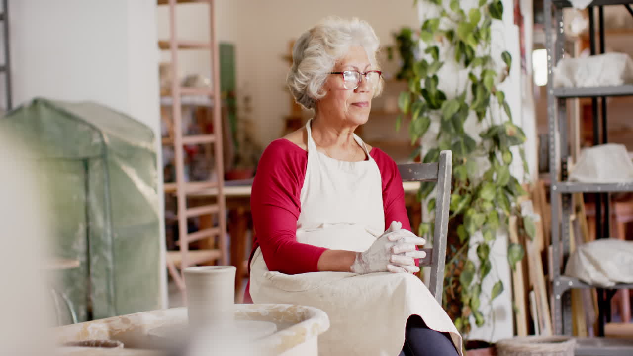 Felice anziana vasaia birazziale con capelli grigi che riposa sorridendo in uno studio di ceramica, al rallentatore