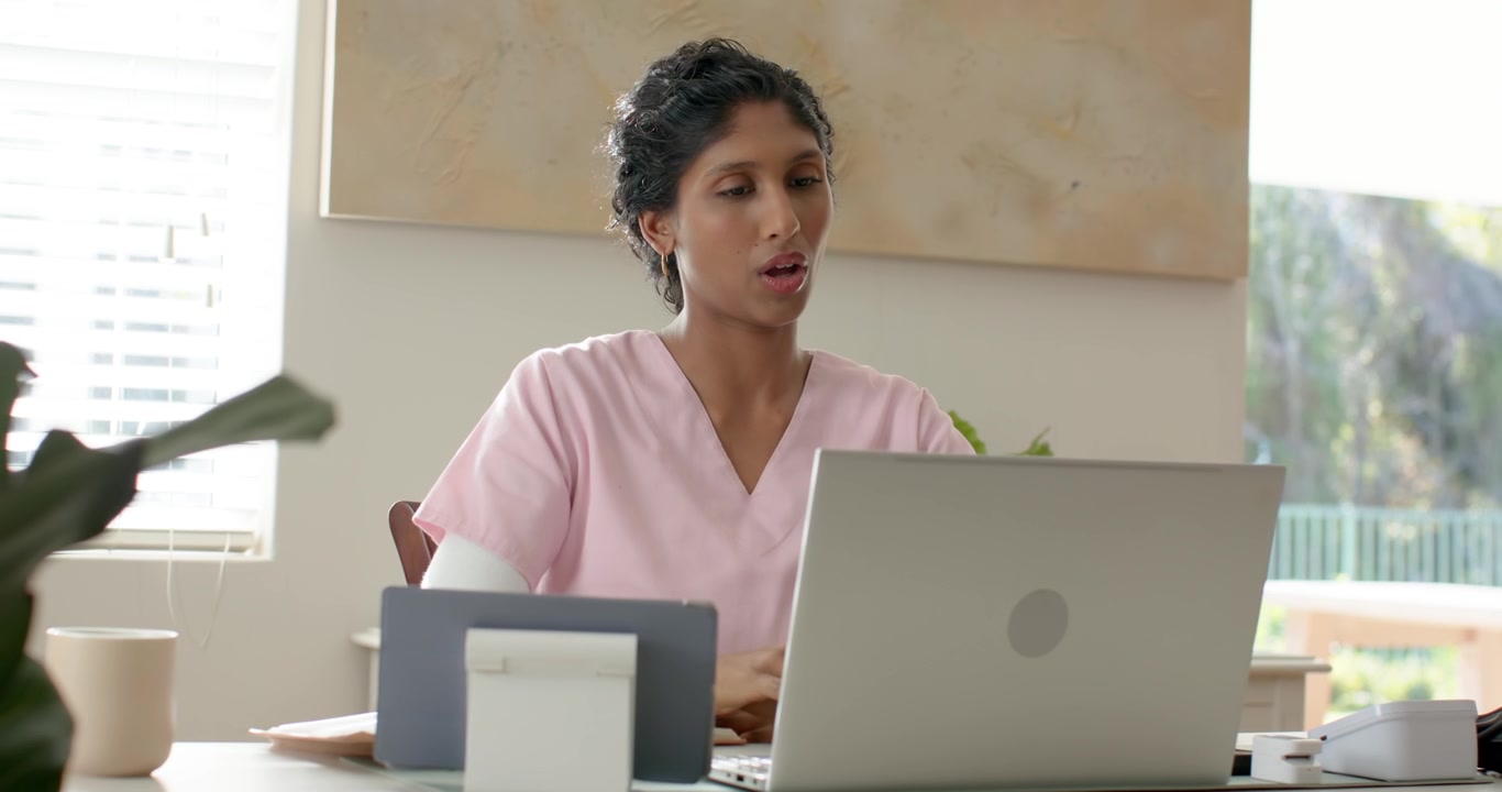Donna afroamericana in camice rosa seduta alla scrivania che osserva un laptop argentato, mentre digita appunti sui pazienti