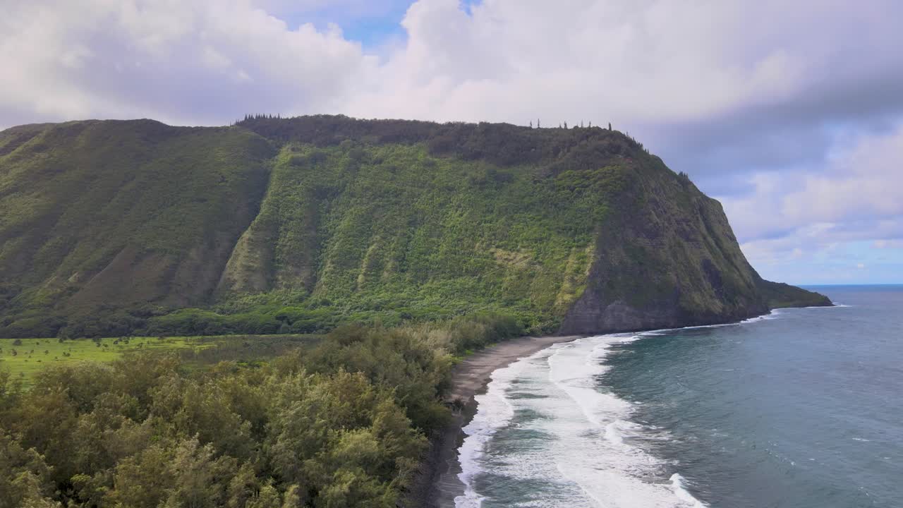 Drone transitions from mountain top to heavy surf on Waipi'o Valley beach under partially cloudy skies on Hawaii's Big Island