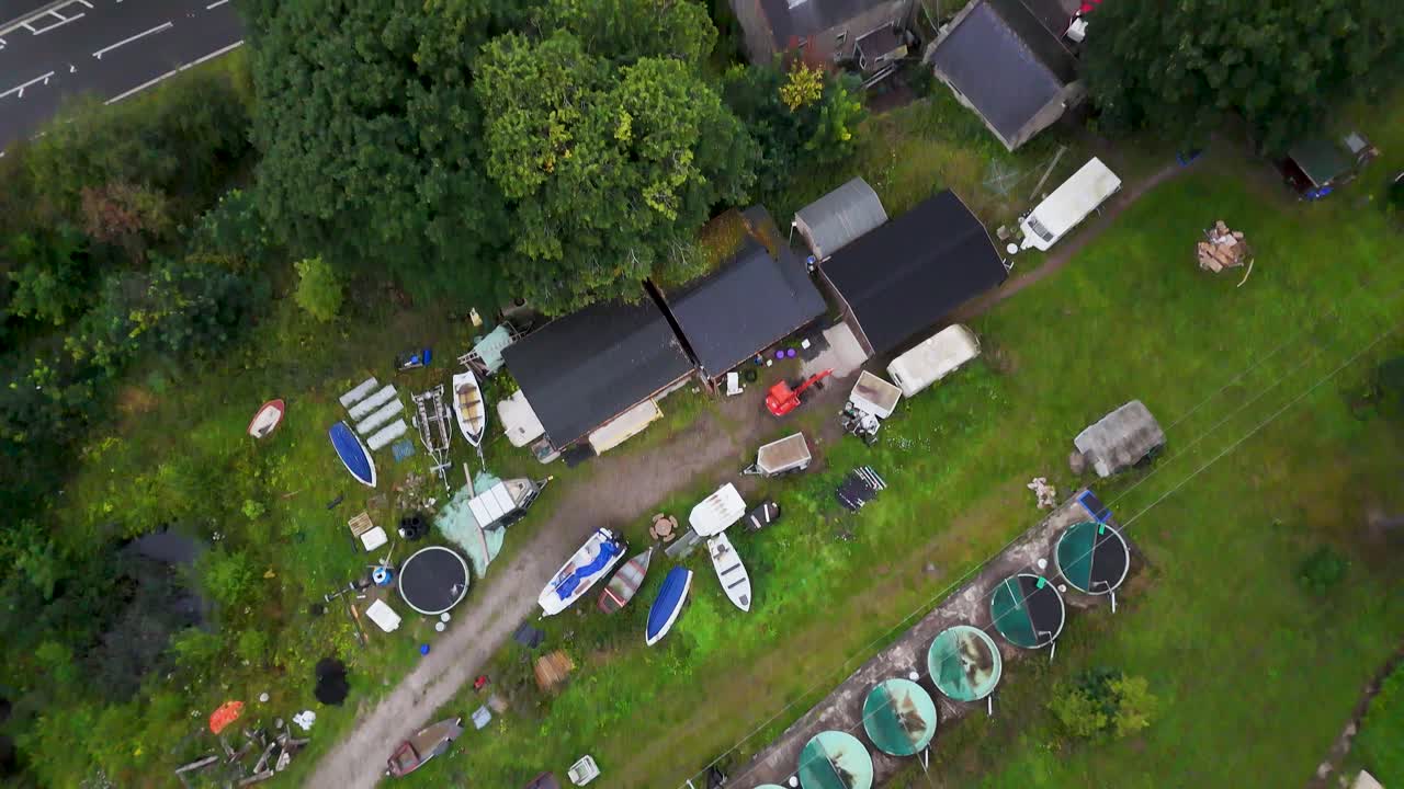 Aerial camera smoothly moves over boathouses, trees, and a road near a reservoir in Hope Valley, England, under natural daylight