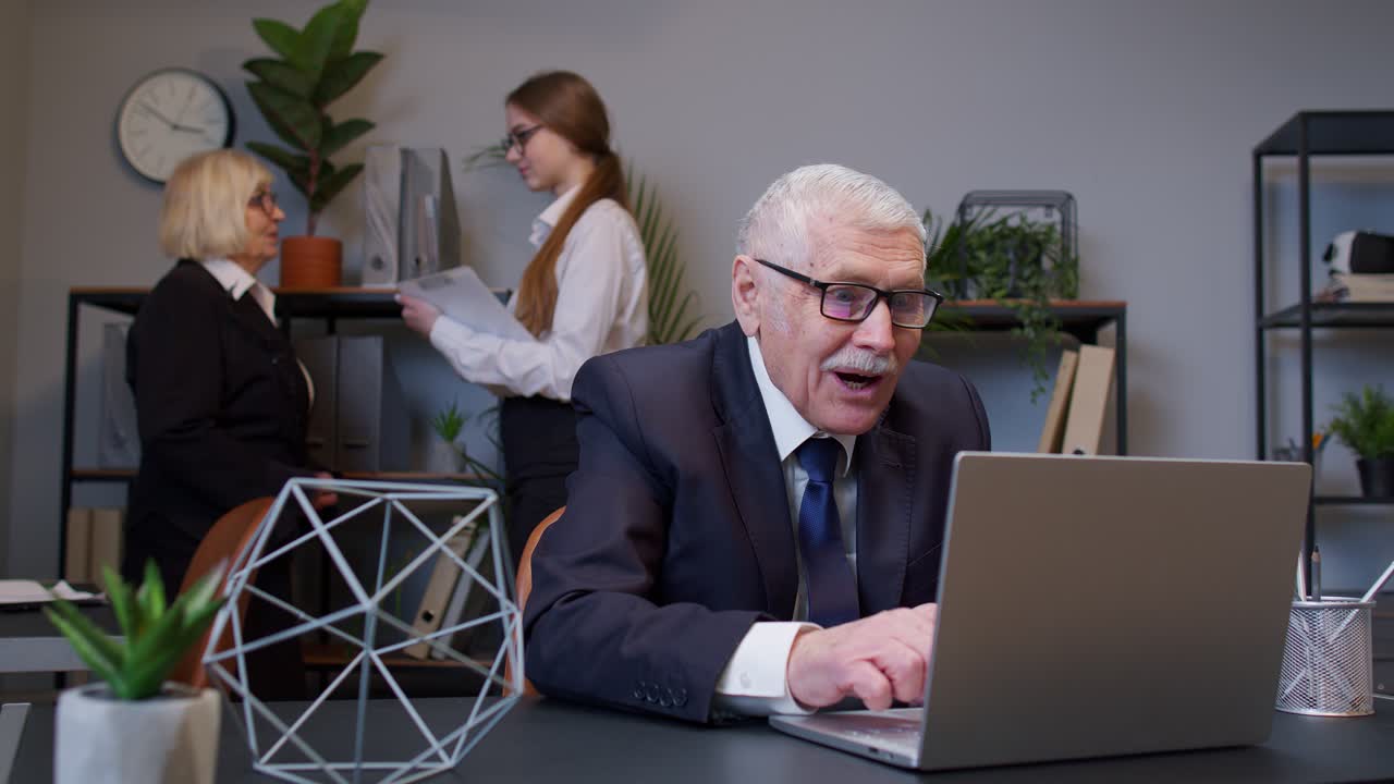 Excited mature old businessman raising hands celebrating sudden victory with colleagues in office