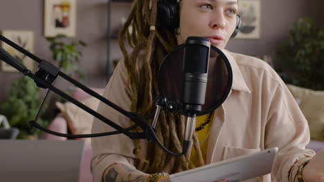 vista de cerca de una mujer con rastas grabando un podcast hablando por un micrófono y sosteniendo una tableta sentada en el escritorio