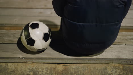 vista posterior de un hombre con una chaqueta negra sentado tranquilamente en un estadio, el hombre está sentado solo con las manos entre las piernas, y una pelota de fútbol se coloca cerca de él en el banco