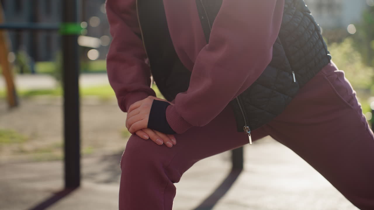 Urban woman in hoodie and quilted vest grips iron rod during forward lunge on urban park tile under sunny sky casting long shadows while focused on form and strength in modern floral environment