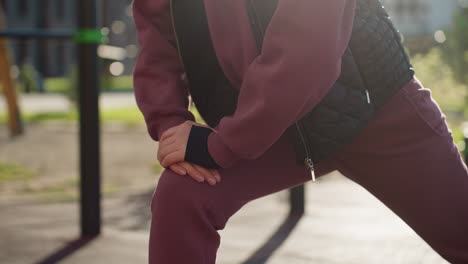 urban woman in hoodie and quilted vest grips iron rod during forward lunge on urban park tile under sunny sky casting long shadows while focused on form and strength in modern floral environment