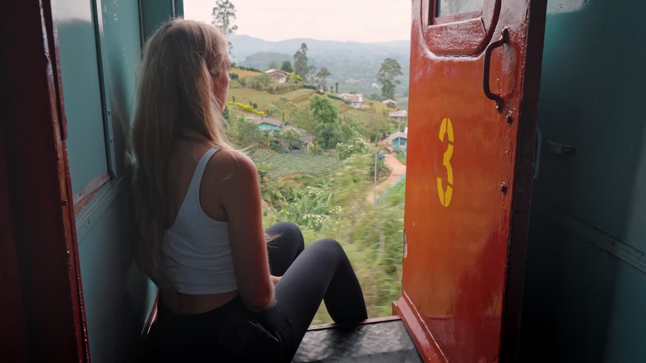 A woman sits peacefully on the open door of a moving train along the iconic Ella to Kandy route in Sri Lanka, taking in the breathtaking views of green hills, tea plantations, and dense jungle.