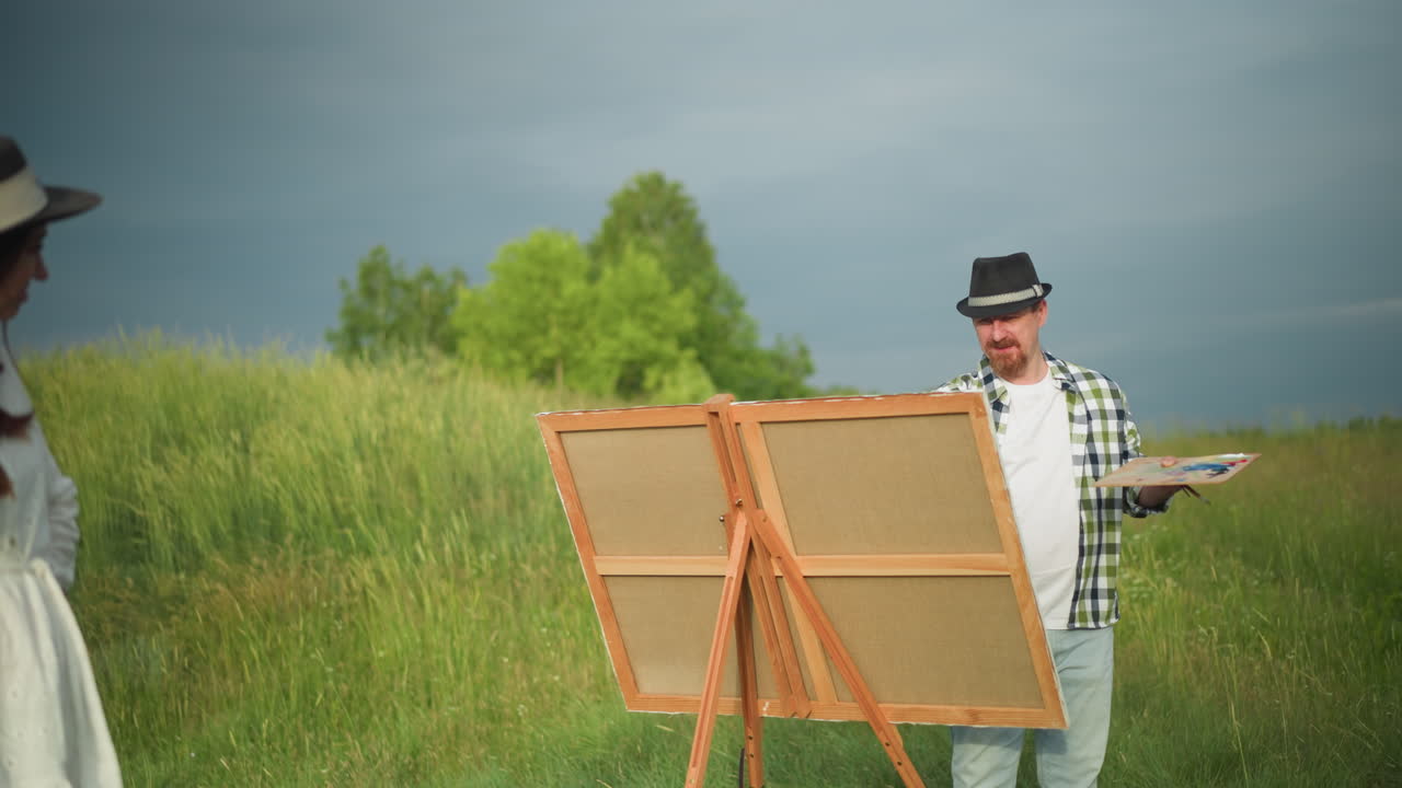 A painter holding a palette and brush captures the image of a woman in a white dress and hat, standing poised in front of him. Set in a lush grassy environment